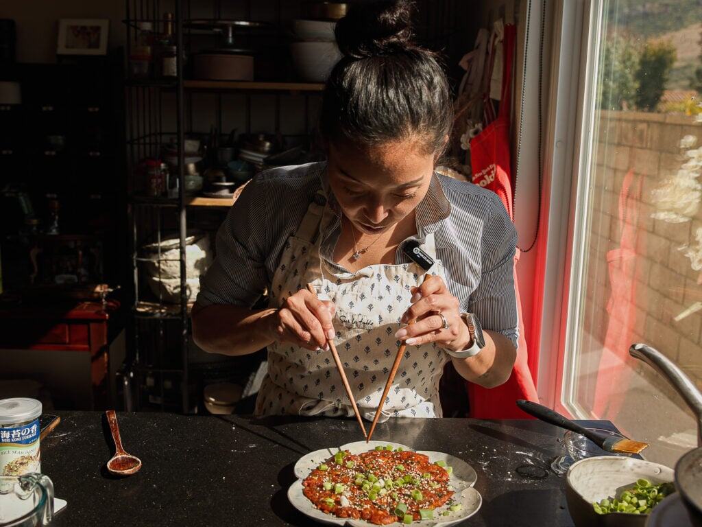 A woman wearing an apron uses chopsticks to garnish a dish with green onions in a sunlit kitchen, standing by a counter near a window.